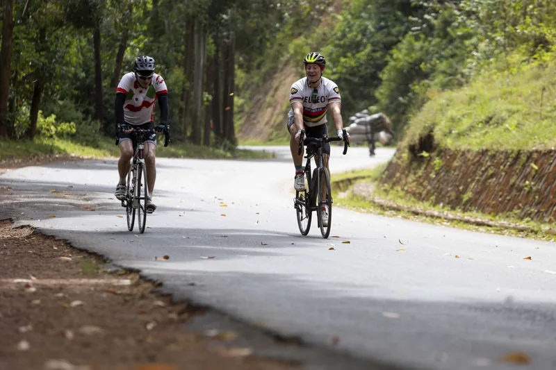 Two riders on a tarmac road walled in by trees, Day 1